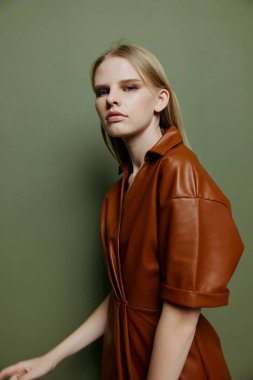 Pankart tasarımı için şık bir yakın plan portresi. Deri kahverengi palet elbiseli seksi sarışın kadın yeşil stüdyo arka planında izole edilmiş bir şekilde kameraya bakıyor.