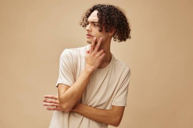 Mutsuz, hoşnutsuz, bronz tenli, tiksindirici tişörtlü harika kıvırcık adam bej arka planda yalnız poz veriyor. Yeni Moda Koleksiyonu teklifi. Halkın Duyguları konsepti. Reklam için boş yer