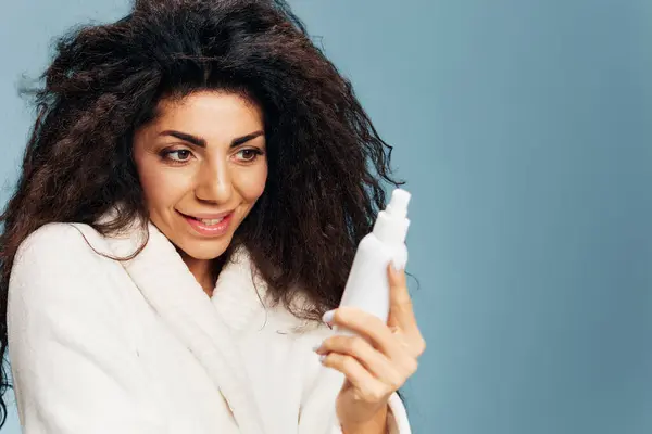 Happy excited curly Latin lady in bathrobe reading ingredients of toner spray in hands posing isolated on pastel blue background, feeling excited of effective skincare routine. Skincare concept