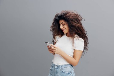 Gadget Bağımlılık Konsepti. Beyaz tişörtlü mutlu kıvırcık kadın cep telefonu kullanıyor, mesaj yazıyor, arkadaşlarıyla sohbet ediyor gri stüdyo arka planında mobil oyun oynuyor.