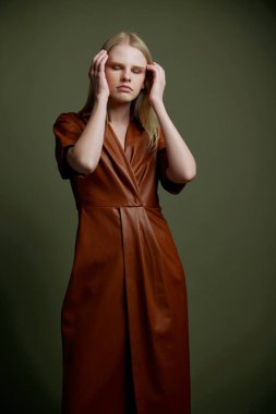 Havalı, şık, kahverengi trençkotlu zarif, kendine güvenen sarışın bayan stüdyo yeşil duvar arka planında el ele tutuşmuş saçlara dokunuyor. Moda bayrağı