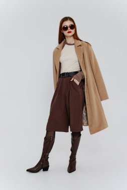 Kırmızı saçlı, gözlüklü ve bej ceketli zarif bir bayan, bir sergi odası reklamı için stüdyoda beyaz bir arka planda poz veriyor. Markalar için harika bir teklif. Yüksek kalite fotoğraf