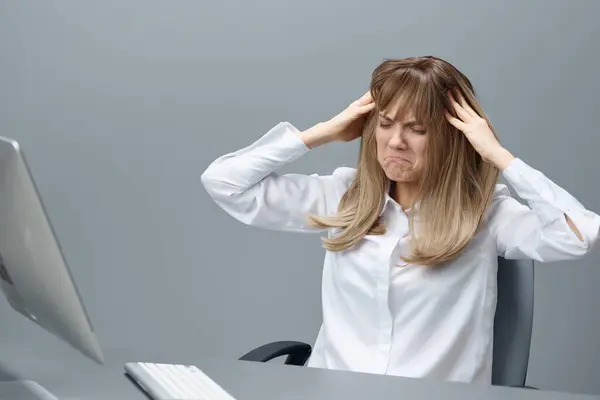 Unhappy pretty blonde businesswoman worker touch head both hands think of problem solution sitting at workplace in gray modern office. Employee work on computer online making big mistake. Copy space