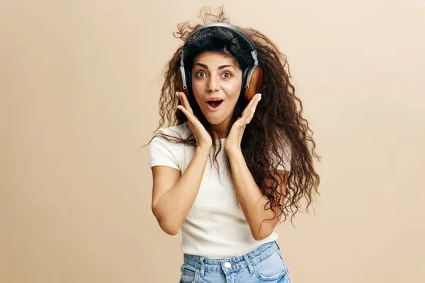 Wow offer. Excited Latin female with afro enjoy cool playlist in wireless earpieces, say Yeah, staying isolated near beige wall. Copy free space for design ad. Fashion IT Seasonal Sale concept