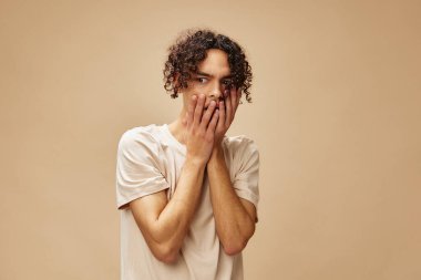 Temel tişörtlü harika bronzlaşmış kıvırcık adam, bej renkli pastel arka planda izole edilmiş pozlar veren yanaklara dokunuyor. Yeni Moda Koleksiyonu teklifi. İnsanlar ve Duygular konsepti. Reklam için boş yer