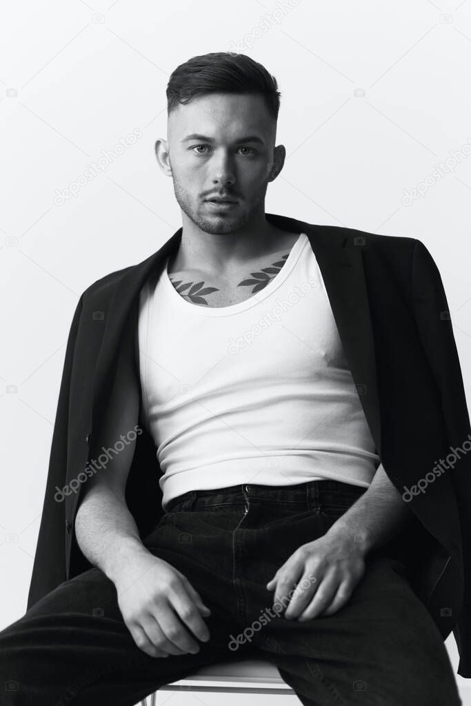 Modelling snapshots. Serious self-confident attractive handsome man in black jacket sitting on chair posing in white studio background. Black and White concept. Copy space
