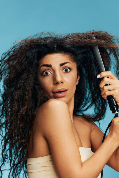 Shocked pretty Latin lady with curly hair using straightener, open mouth, posing isolated on blue wall background. Hair routine concept, haircare, hair ironing, hairdressing, dry damaged hair