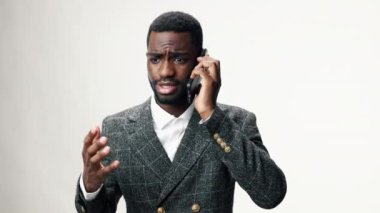 Afro-Amerikalı bir işadamı telefonda sinirli ve agresif bir şekilde konuşuyor iş elbisesi giymiş gri bir duvara karşı ve elleriyle işaret ediyor. Yüksek kalite 4k görüntü