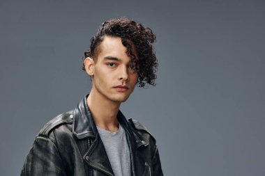 Ciddi bir şekilde bronzlaşmış kıvırcık deri ceketli adam portresi gri stüdyo arka planında izole edilmiş kameraya bakıyor. Güzel moda teklifi. Büyük Mevsimlik Satış Yeni Koleksiyon konsepti. İlan için alanı kopyala
