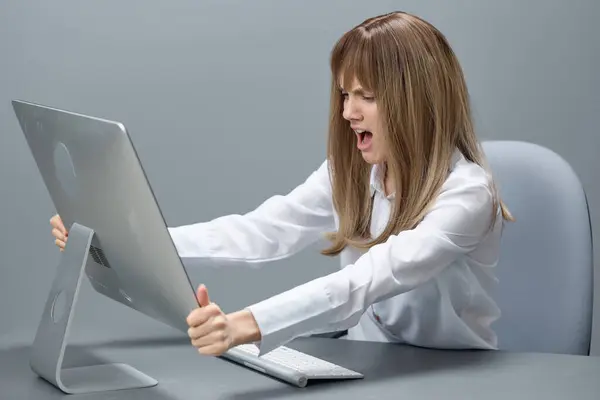 Angry evil pretty blonde businesswoman screaming to desktop Raging after computer software crash error sitting at workplace in gray modern office. Technology And Operation Problem Concept. Copy space