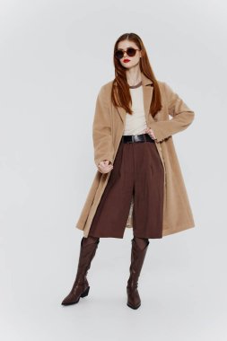 Kırmızı saçlı, gözlüklü ve bej ceketli zarif bir bayan, bir sergi odası reklamı için stüdyoda beyaz bir arka planda poz veriyor. Markalar için harika bir teklif. Yüksek kalite fotoğraf