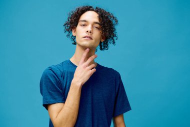 Kafası karışmış, komik kıvırcık, bronzlaşmış, temel tişörtlü beyaz adam mavi arka planda yalnız poz veriyor. Yaşam tarzı ve Duygular konsepti. Reklam için fotokopi alanı için iyi teklif.