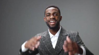 Afro-Amerikalı bir adam sırtını kameraya çevirir ve ellerini zevk ve mutluluk içinde kaldırır, gri bir arka planda güzel bir ceket ve beyaz bir gömlek giyer. Yüksek kalite 4k görüntü