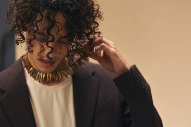 Klasik kahverengi ceketli harika bronzlaşmış kıvırcık adam bej renkli pastel arka planda izole edilmiş etnik kolyeye dokunuyor. Yeni Moda Koleksiyonu teklifi. Retro stil konsepti. Reklam için boş yer