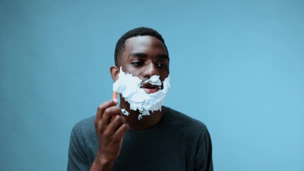 アフリカ系アメリカ人男性は カミソリでひげを剃り 青い背景にあるTシャツに微笑みます 高品質の4K映像 — ストック動画