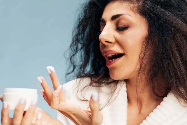 Closeup portrait of pretty curly Latin lady in bathrobe apply organic day cream perform morning skincare beauty procedures posing isolated on pastel blue background. Cosmetic product ad concept