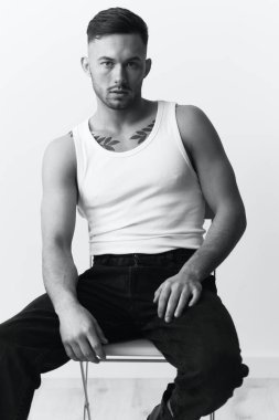 Fotoğrafları modelliyorum. Kendine güveni tam, beyaz fanilalı yakışıklı beyaz bir adam beyaz stüdyo arka planında sandalyede poz veriyor. Siyah ve Beyaz konsepti. Boşluğu kopyala