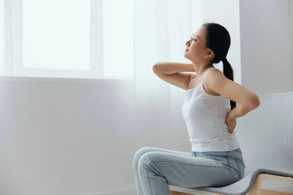 Back pain. Suffering from osteochondrosis after long study work pretty young Asian woman touching painful lower back at home interior living room. Injuries Poor health Illness concept. Cool offer