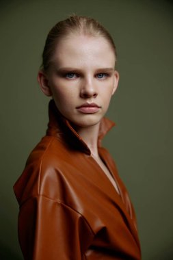 Şık, ciddi, mavi gözlü, gece makyajlı, deri elbiseli genç bir kız izole bir şekilde poz veren kameraya bakıyor. Yeşil arka planda stüdyo portresi. Moda.