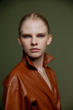 Şık, ciddi, mavi gözlü, gece makyajlı, deri elbiseli genç bir kız izole bir şekilde poz veren kameraya bakıyor. Yeşil arka planda stüdyo portresi. Moda.