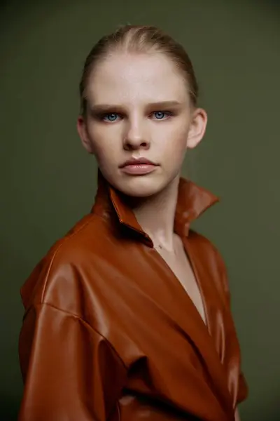 Stylish serious young blonde girl with blue eyes with evening makeup in a leather dress looks at the camera posing isolated. Studio portrait on a green background. Fashion