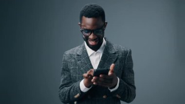 Elinde telefon tutan Afrikalı Amerikalı bir işadamı ona bakıyor ve gri bir arkaplanda kazanmaktan ve kazanmaktan mutlu oluyor. Yüksek kalite 4k görüntü