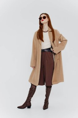 Kırmızı saçlı, gözlüklü ve bej ceketli zarif bir bayan, bir sergi odası reklamı için stüdyoda beyaz bir arka planda poz veriyor. Markalar için harika bir teklif. Yüksek kalite fotoğraf