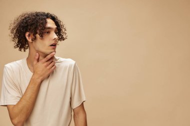 Esnek tişörtlü bronzlaşmış harika kıvırcık adam bej renkli pastel arka planda yalnız poz veriyor. Yeni Moda Koleksiyonu teklifi. İnsanlar ve Duygular konsepti. Reklam için boş yer