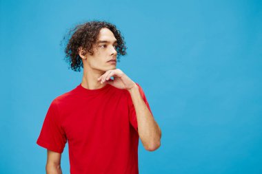 Düşünceli, düşünceli, mutsuz, bronzlaşmış, beyaz tenli, temel kırmızı tişörtlü adam mavi arka planda yalnız poz veriyor. Yaşam tarzı ve Duygular konsepti. Reklam için fotokopi alanı için iyi teklif.