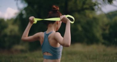 Genç bir kadın spor giyim kuşamında parkta spor yapıyor ve sırtını sıvazlıyor. Lastik bir bantla egzersiz yapmak. Yüksek kalite 4k görüntü