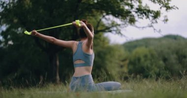 Genç bir kadın spor giyim kuşamında parkta spor yapıyor ve sırtını sıvazlıyor. Lastik bir bantla egzersiz yapmak. Yüksek kalite 4k görüntü