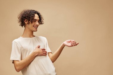 Temel tişörtlü, bronzlaşmış komik kıvırcık adam bej renkli pastel arka planda yalnız poz veriyor. Yeni Moda Koleksiyonu teklifi. İnsanlar ve Duygular konsepti. Reklam için boş yer