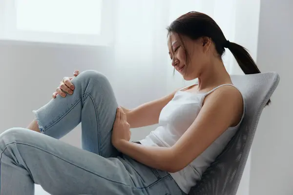 Unhappy sad suffering tanned beautiful young Asian woman touch injury from training knee at home interior living room. Injuries Poor health Illness Broken Bone in Leg concept. Cool offer Banner