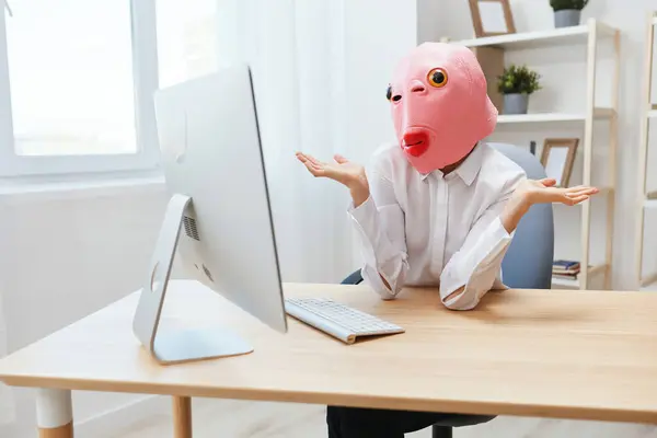 Pensive crazy businesswoman worker freelancer in pink fish mask work on computer online pondering or making decision thinking of problem solution spread hands in light modern office. Copy space