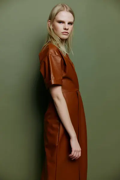 Self-confident fashionable blonde young model looking over her shoulder at camera dress in brown leather trench coat posing isolated on over olive green studio wall background. Stylish offer