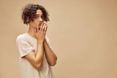 Temel tişörtlü harika bronzlaşmış kıvırcık adam, bej renkli pastel arka planda izole edilmiş pozlar veren yanaklara dokunuyor. Yeni Moda Koleksiyonu teklifi. İnsanlar ve Duygular konsepti. Reklam için boş yer