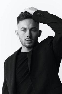 Fotoğrafları modelliyorum. Kendine güvenen, ceketli yakışıklı kaslı adamın yakın plan portresi beyaz stüdyo arka planında oturan sandalyeye bakıyor. Siyah ve Beyaz konsepti. Boşluğu kopyala