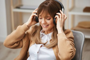 Hoş, mutlu, genç, sarışın, serbest çalışan, kalın kazaklı bayan kulaklık takıyor ve modern iç mekandaki koltukta oturan favori şarkıların tadını çıkarıyor. Müzik Zamanı Rahatlatıcı Havalı Parça Listesi Konsepti. Boşluğu kopyala