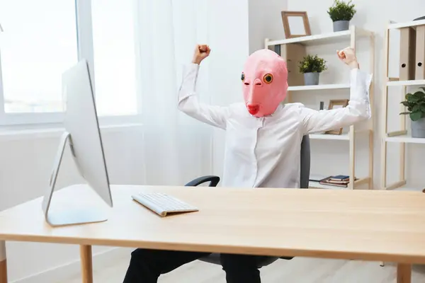 穿着粉色鱼面具的疯狂而快乐的女自由职业者在网上电脑上工作 展示了轻松现代办公室里的肌肉 复制空间 — 图库照片