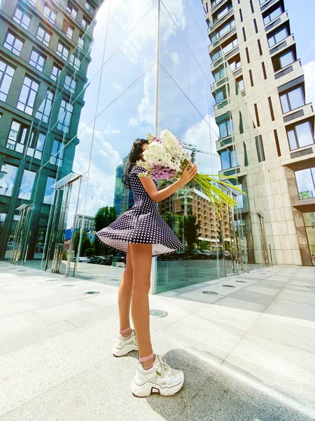 在这座城市的摩天大楼前 穿着飘扬的短裙和白色运动鞋的年轻模特站在户外 手里拿着一束白花和紫花 全长照片 — 图库照片