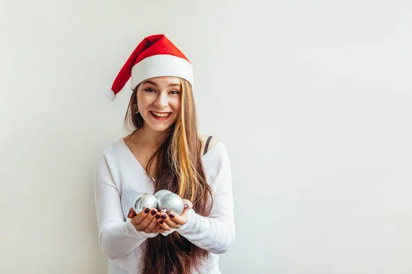 戴着红色圣诞老人帽的漂亮姑娘拿着圣诞装饰球 被白色的背景隔开 看上去既高兴又兴奋 年轻女人的肖像 真实的情感 圣诞佳节快乐 — 图库照片