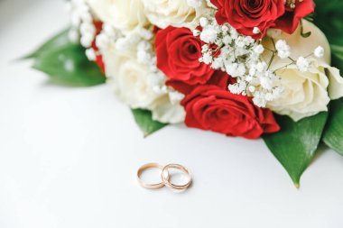 Beyaz arka planda duran altın alyanslar ve çiçek buketi beyaz kırmızı renklerde. Aşk ilanı, bahar. Düğün kartı, Sevgililer Günü tebriği. Evlilik yüzükleri. Düğün günü detayları