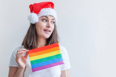 Kırmızı Noel Baba şapkalı güzel lezbiyen kız beyaz arka planda izole edilmiş LGBT gökkuşağı bayrağı ile mutlu ve heyecanlı görünüyor. Genç kadın Gay Gurur portresi. Mutlu Noeller ve yeni yıl tatili.