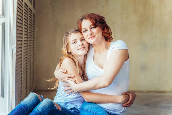 Jeune mère embrassant son enfant. Femme et adolescente se détendre dans une chambre blanche près d'un mur gris à l'intérieur. Bonne famille à la maison. Jeune maman jouant avec sa fille. — Photo