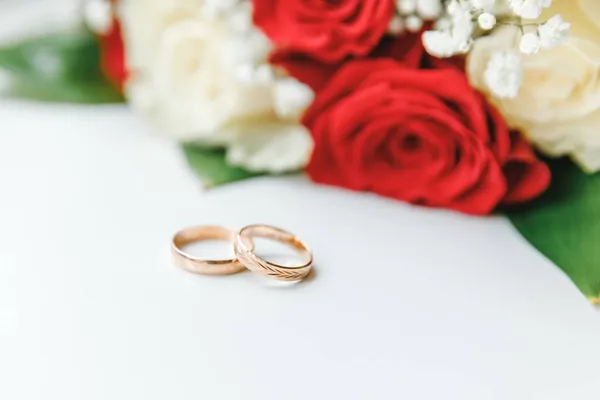Arany jegygyűrűk és virágok csokor fehér piros színekben fekvő fehér háttér. Szerelmi nyilatkozat, tavasz. Esküvői kártya, Valentin napi üdvözlés. Jegygyűrűk. Esküvő napja részletek. — Stock Fotó