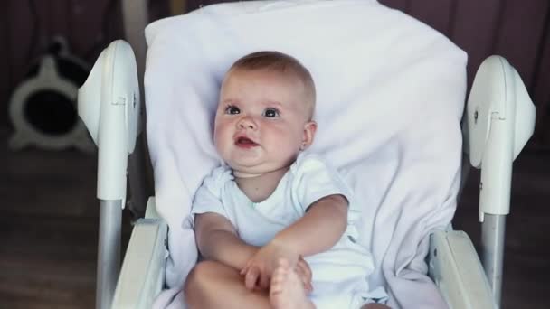 Aranyos, mosolygó arcú újszülött lány, aki fehér háttérrel nézi a kamerát. Csecsemőbaba pihen, otthon fekszik az etetőszéken. Anyaság boldog gyermek koncepció. — Stock videók
