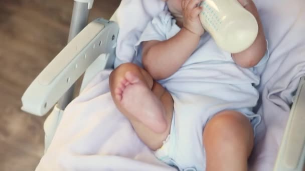 Χαριτωμένο μικρό νεογέννητο κορίτσι πίνοντας γάλα από το μπουκάλι και κοιτάζοντας κάμερα σε λευκό φόντο. Βρέφος θηλάζει τρώει τη διατροφή του γάλακτος ξαπλωμένος στην καρέκλα σίτισης στο σπίτι. Μητρότητα ευτυχισμένο παιδί — Αρχείο Βίντεο