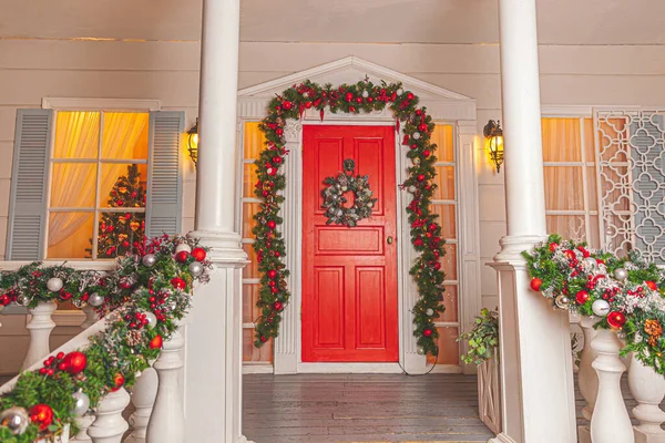 Idéia da decoração do alpendre do Natal. Entrada da casa com porta vermelha decorada para férias. grinalda grinalda vermelha e verde de ramos de abeto e luzes sobre trilhos. Véspera de Natal em casa — Fotografia de Stock