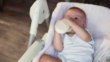 Şişeden süt içen ve beyaz arka planda kameraya bakan tatlı küçük bir kız. Bebek emzirme sütü yiyor ve evdeki beslenme sandalyesine uzanıyor. Annelik mutlu çocuk