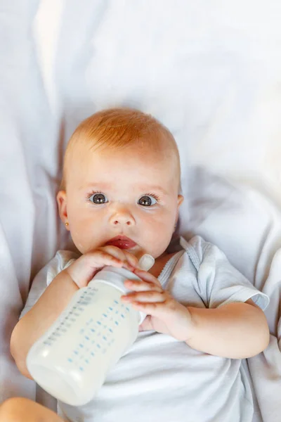Aranyos újszülött lány tejet iszik üvegből, és fehér háttérrel nézi a kamerát. A csecsemő csecsemő szopogatja a tejet, miközben otthon fekszik az ágyon. Anyaság boldog gyermek koncepció. — Stock Fotó